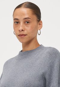 Light gray sweater with a high neckline, ribbed texture, and a relaxed fit. Model wears silver hoop earrings.
