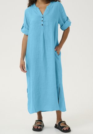 Kobieta w luźnej, jasnoniebieskiej sukience midi na guziki z podwiniętymi rękawami oraz czarnych wsuwanych sandałach z ozdobnym przeszyciem.