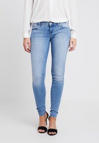 Vaqueros ajustados de un azul claro, confeccionados en denim, con un diseño clásico de cinco bolsillos y una textura suave. Llevados con sandalias negras.