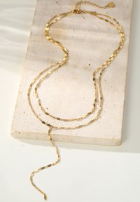 Goldfarbene, mehrschichtige Halskette mit einer zarten Kette mit unregelmäßigen Gliedern und einem verstellbaren Verschluss, präsentiert auf einer texturierten Steinfoberfläche.