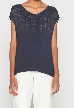 Femme portant un haut bleu marine à manches courtes avec des motifs ponctués verticaux argentés et un pantalon blanc, montrée des épaules aux hanches.