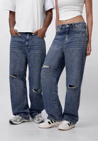 Light wash denim jeans with a loose fit, featuring distressed rips at the knees. Paired with white t-shirts and casual sneakers.