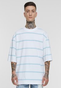 Tricou alb supradimensionat cu dungi orizontale de culoare albastră deschisă. Material din bumbac, gât rotund, mâneci scurte și croială lejeră. Tatuaj distinct vizibil.