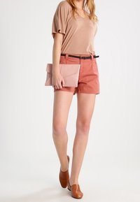 Light brown top, coral shorts with a black belt, holding a pink envelope clutch, and wearing brown slip-on shoes. Smooth textures throughout.