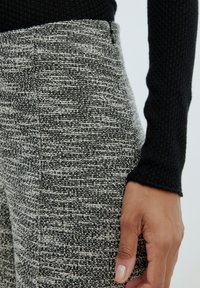 Strukturierter schwarzer und weißer Stoff von maßgeschneiderten Hosen, mit sichtbaren Nähten und einem seitlichen Reißverschluss. Ein Ärmel eines schwarzen Pullovers ist teilweise sichtbar.