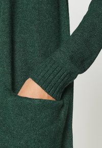 Vihreä neulepaita, jossa on resoritukikehys ja sivutasku. Kangas näyttää pehmeältä ja siinä on teksturoitu pinta.