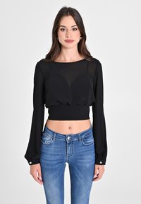 Schwarzes, kurzes Langarmoberteil aus leichtem Stoff mit gerafftem Vorderteil. Kombiniert mit blauen, gut sitzenden Jeans mit heller Ausbleichung und Taschen.