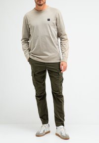 Helles beige Langarmhemd mit kleinem schwarzem Logo, kombiniert mit olivgrünen Cargo-Hosen und weißen Sneakern mit grauen Akzenten.