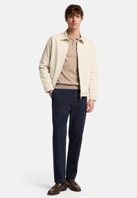 Beige Bomberjacke, hellbraunes Poloshirt, marineblaue Hose, braune Schuhe. Der Stoff wirkt glatt; die Jacke verfügt über einen vorderen Reißverschluss und einen Kragen.