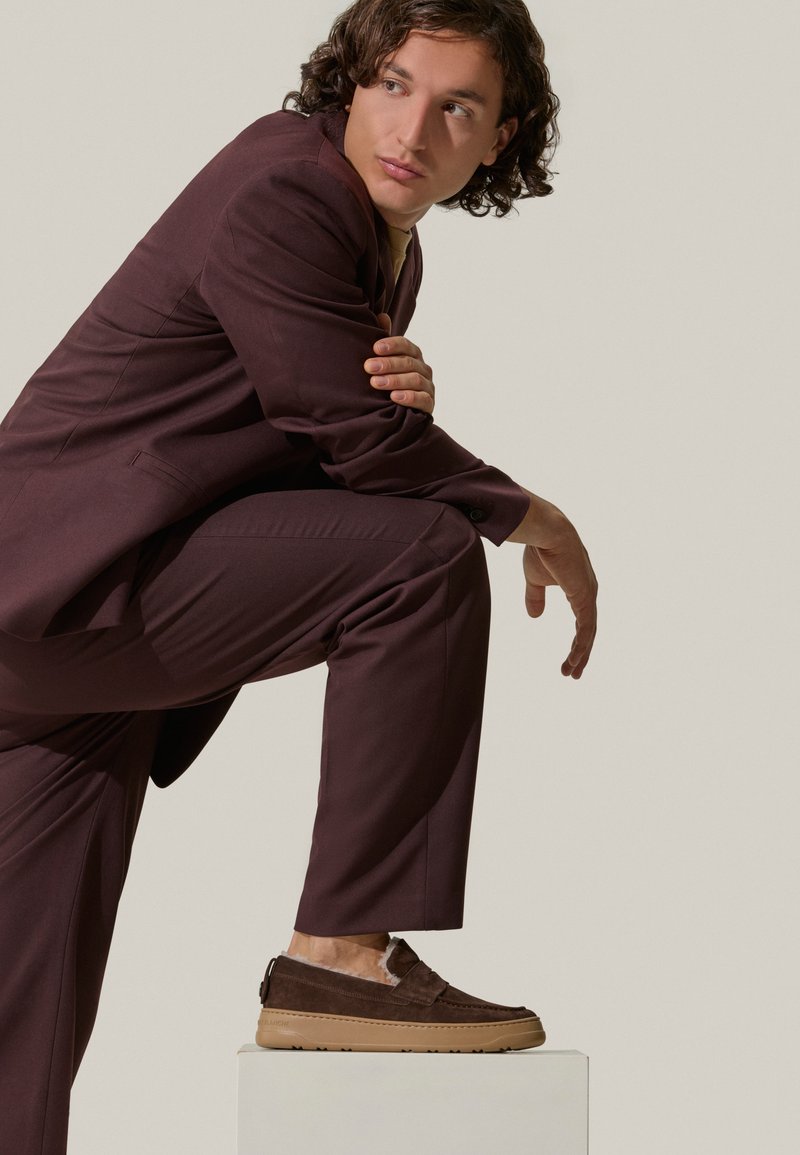 Brown suede slip-on shoes with beige rubber soles, textured accents, worn with a deep burgundy suit. The subject poses on a white pedestal.