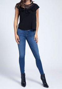Blusa negra con detalles de encaje y volantes en el escote, combinada con jeans ajustados azules y botines negros con diseño de punta.