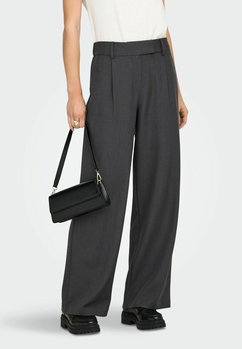 Personne portant un pantalon large gris foncé, un haut blanc, des mocassins noirs épais, tenant un petit sac à bandoulière noir à côté d'elle.