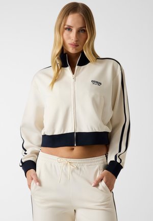 Femme portant un survêtement crème zippé avec des bandes noires et le logo "Guess", les mains dans les poches, debout sur un fond uni.