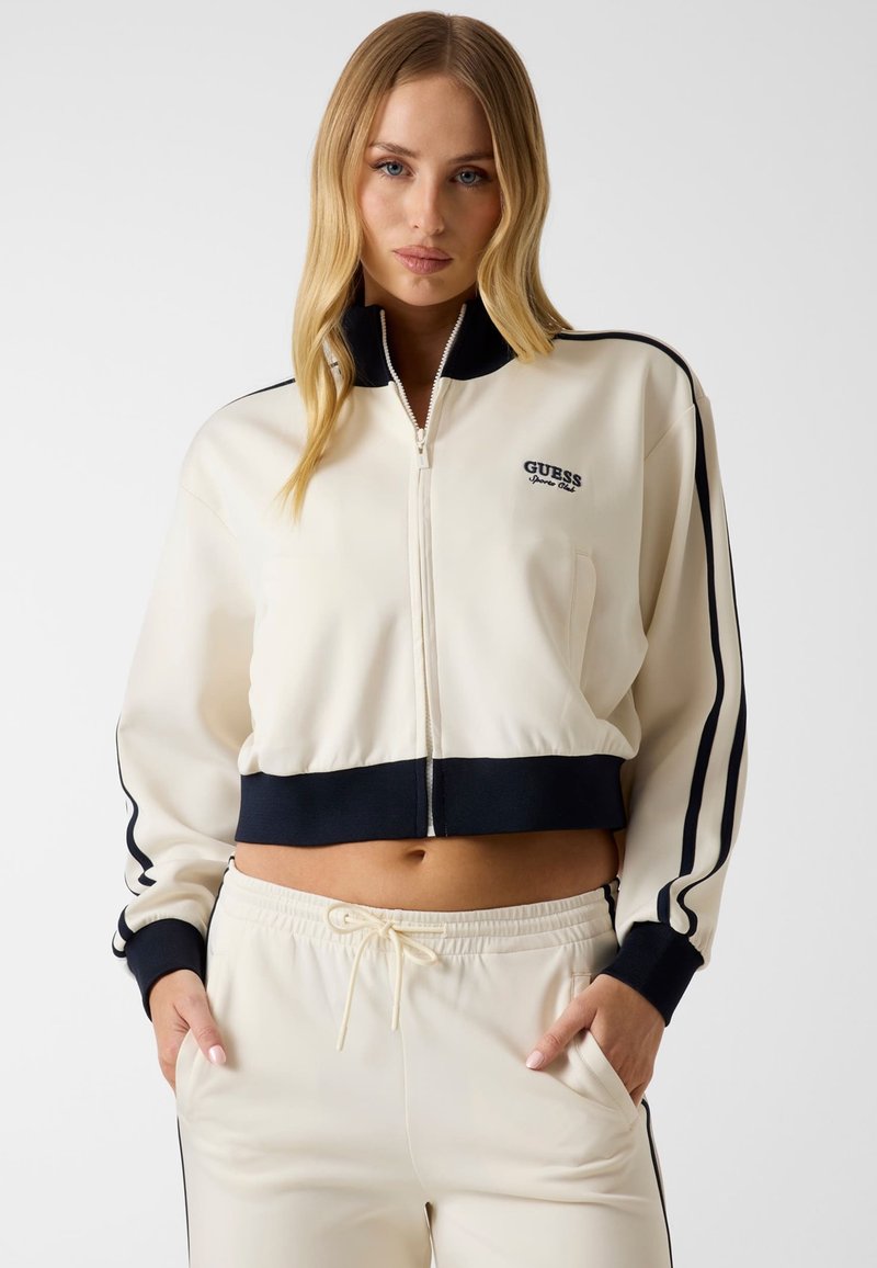 Femme portant un survêtement crème zippé avec des bandes noires et le logo "Guess", les mains dans les poches, debout sur un fond uni.