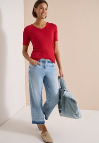 Femme souriante portant un haut rouge à manches courtes, un jean large bleu clair, des chaussures beiges type slip-on, portant un grand sac fourre-tout en denim, debout dans un espace neutre.