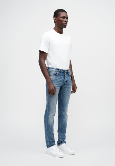 Balta trumpa rankovėmis marškinėliai derinami su šviesiai mėlynomis džinsomis, turinčiomis liekną siluetą ir švelnų blukimą, papildyti baltais sportbačiais.