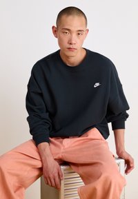 Fekete pulóver, kerek nyakkivágással és hosszú ujjakkal, fehér Nike логóval. Világos rózsaszín nadrággal viselve, egyszínű háttér előtt.