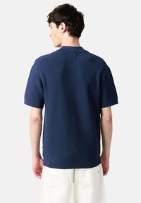 Marinblå kortärmad skjorta med en texturerad stickad design, med en platt krage och sidofrekvenser, bärs ihop med krämfärgade byxor.