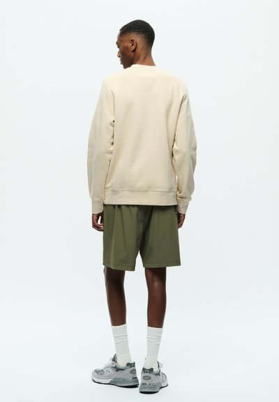 Beige Sweatshirt aus strukturiertem Stoff, lockere Passform. Olivengrüne Shorts, knielang. Graue Sneakers mit Akzenten und weißen Socken. Neutraler Hintergrund.