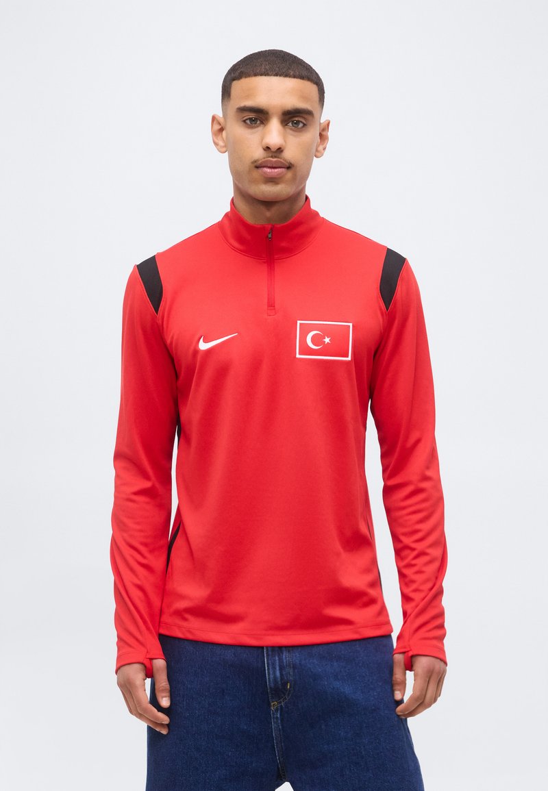 Άνδρας που φοράει κόκκινο αθλητικό μπουφάν Nike με σχέδιο της τουρκικής σημαίας, στέκεται μπροστά σε απλό ανοιχτό γκρι φόντο.