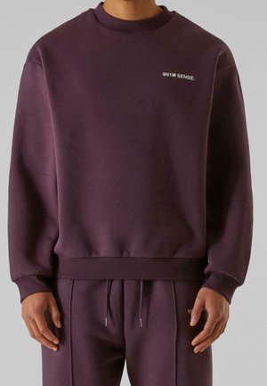 Homme portant un sweat-shirt violet foncé et un pantalon assorti avec un petit texte blanc « 9N1M SENSE. » sur la poitrine et la ceinture du pantalon.