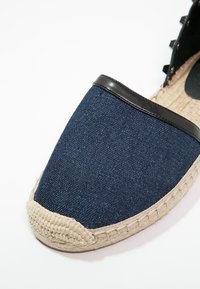 Tummansininen espadrille, jossa on denimpintainen päällinen, musta nahkareunus ja kudottu juuttipohja. Ominaisuuksiin kuuluu sileä pinta ja pyöreä kärjellinen muoto.