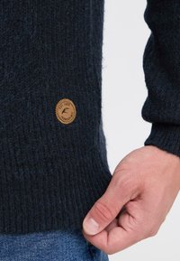 Gebreide donkerblauwe trui met ribbelstructuur, voorzien van een ronde bruine leren patch met een reliëflogo aan de rand.