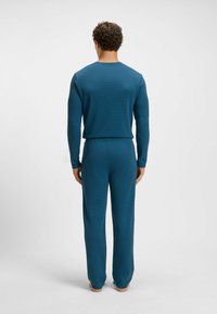 Homme aux cheveux bouclés se tient pieds nus, de dos, portant un ensemble coordonné en haut et en bas à manches longues en bleu sarcelle, sur un fond uni.