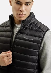 Czarna kamizelka puchowa z poziomym pikowaniem, zamek z przodu i detal z logo na klatce piersiowej, noszona na szarym swetrze.