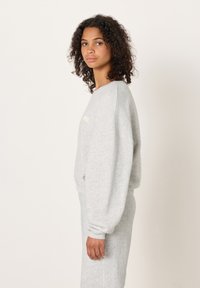 Siva majica s okruglim izrezom, labavog kroja i dugim rukavima. Mekani materijal s minimalnim šavovima, sa suptilnim izvezenim detaljima.