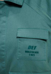 Grüner Stoff mit glatter Textur verfügt über eine Brusttasche. Aufgedruckter Text lautet "DEF WORKATION MODE F/W23" in kontrastierendem Dunkelgrün.