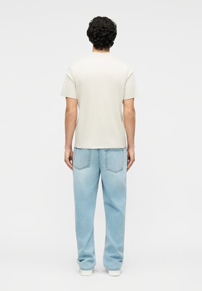 Άνδρας με σκούρα σγουρά μαλλιά, που φοράει ένα απλό ανοιχτό μπεζ κοντομάνικο πουκάμισο και χαλαρά ανοιχτό μπλε τζιν, στέκεται γυρισμένος προς τα πίσω.