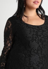 Svart spetsklänning med blommönster, långa genomskinliga ärmar, rundad halslinje och en figursydd överdel med en texturerad överlag.