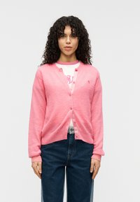 Ροζ μάλλινο ζακέτα με απαλή υφή, V-λαιμόκοψη, κουμπιά εμπρός και μια μικρή λεπτομέρεια με λογότυπο. Φορεμένο πάνω από ένα λευκό πουκάμισο με ροζ λεπτομέρειες.