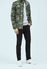 Maastokuvioinen takki pikalukoilla, harmaa collegepaita, mustat slim fit -farkut ja valkoiset lenkkarit punaisilla yksityiskohdilla.