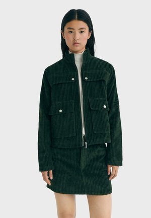 Young woman wearing dark green corduroy jacket with snap pockets and matching skirt, standing against plain light background.