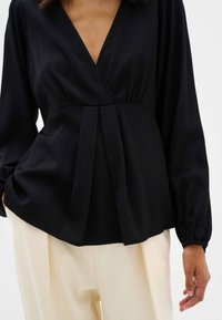 Blusa negra con mangas largas, escote en V, detalle de cintura plisada y una textura suave brillante. Combinada con pantalones de color crema.