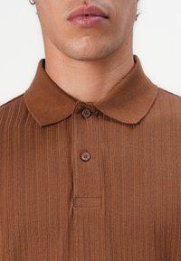 Hnědý žebrovaný polo tričko s knoflíkovým límcem, dvěma knoflíky a jemnou texturou, navržené pro přiléhavý střih.