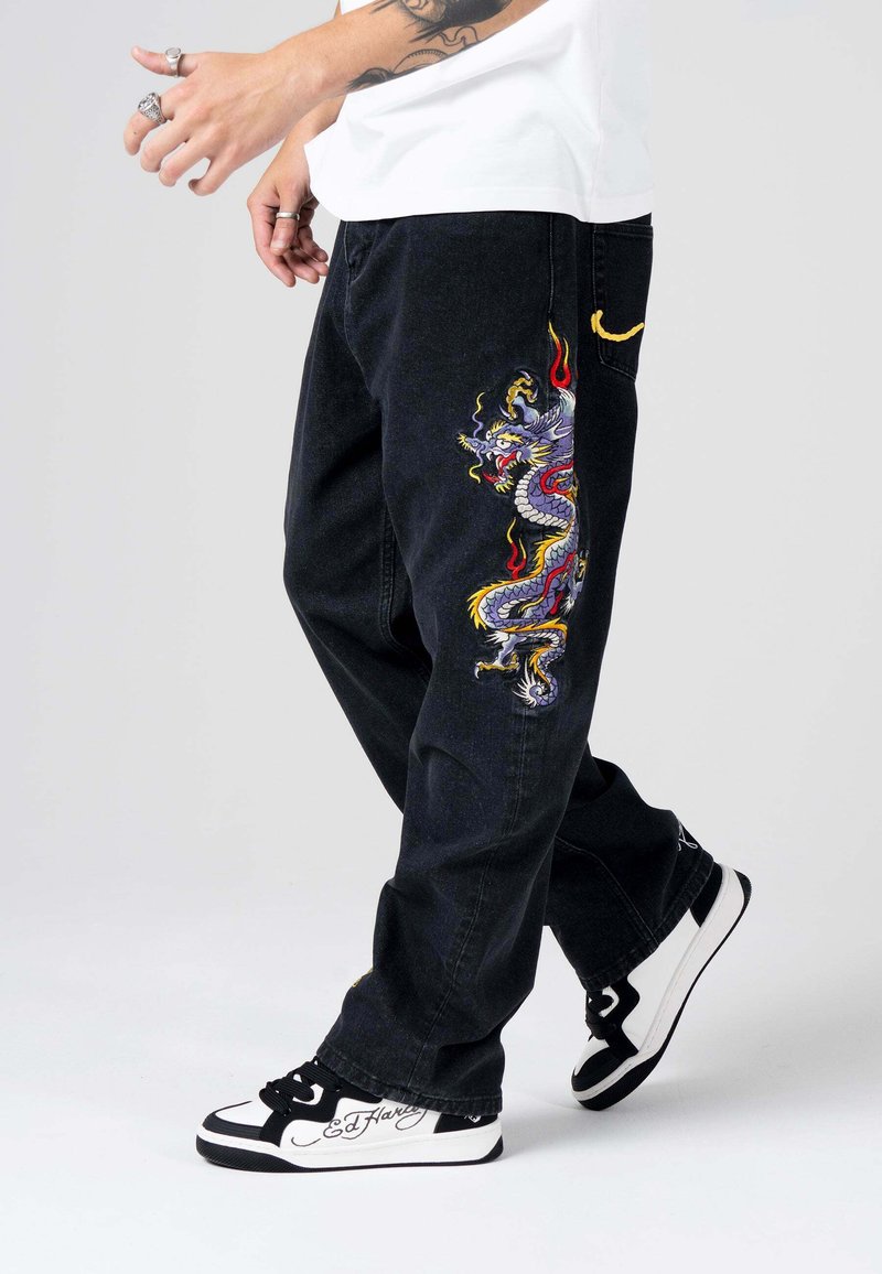 Pantaloni in denim nero con design di drago colorato ricamato sulla gamba sinistra. Presentano una vestibilità ampia e tasche standard, abbinati a sneaker bianche e nere.