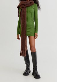 Groene geribde jurk met lange mouwen, een bruine fluffy sjaal om de nek gedrapeerd en kniehoge zwarte leren laarzen.