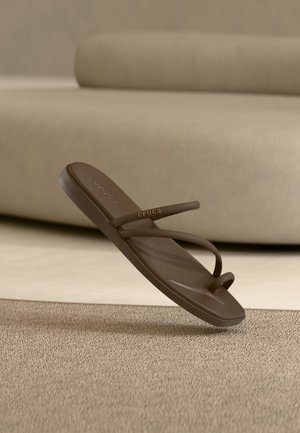 Sandale tong Crocs marron avec double brides et boucle pour l’orteil, survolant un tapis beige texturé avec un canapé beige en arrière-plan.