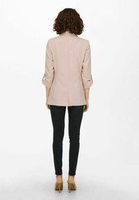 Ljusrosa blazer med rullade ärmar och en springa bak, kombinerad med svarta smala jeans och beige klackar. Slät tygtextur.