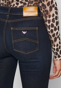 Tumedast sinisest denimist teksased, mille vööl on nahkne Emporio Armani silt, vastanduvad kollased õmblused ja roosa tikandiga logo taskul.