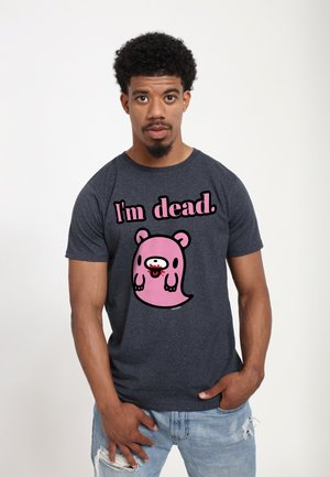 Uomo con t-shirt scura con fantasma rosa dei cartoni animati e testo "Sono morto." In piedi con una mano nella tasca dei jeans azzurri strappati su sfondo bianco.