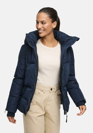 Doudoune marine avec un col haut, un design matelassé et une fermeture éclair sur le devant. Associée à un léger pull et un pantalon beige, affichant une coupe décontractée.