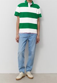 Nuori henkilö, joka on pukeutunut vihreä-valkoiseen raidoitettuun lyhythihaiseen polo-paitaan, vaaleansinisiin farkkuihin ja beessiin tennareihin, seisoo harmaata seinää vasten.