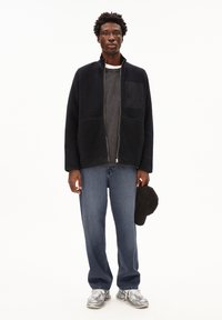 Schwarze Zip-Up-Fleecejacke mit strukturierter Oberfläche, graues T-Shirt darunter, blaue Jeans und graue Sneaker. Halten eine schwarze Baseballmütze.