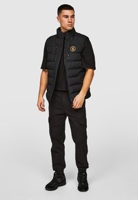 Zwarte gewatteerde vest met hoge kraag, voorzien van een voorrits, twee ritszakken en een rond logo-patch op de borst. Gedragen over een zwart shirt.