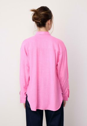 Ženska z lasmi spetimi v figo, oblečena v ohlapno roza majico z dolgimi rokavi in temne hlače, videna od zadaj na enobarvni podlagi.