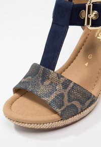 Sandale à bout ouvert avec bride cheville en daim bleu, boucle dorée, et bride avant texturée marron à motifs, sur une semelle beige coussinée.